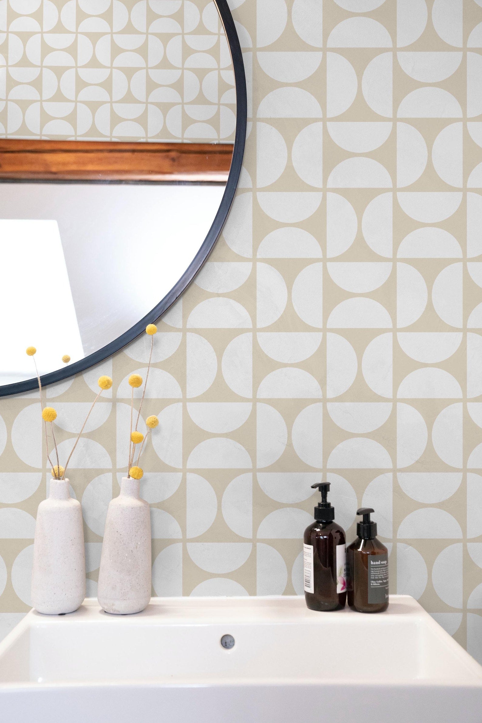 a bathroom setting with a white sink, a round mirror, and a shelf with two bottles of soap and two white vases with yellow flowers.