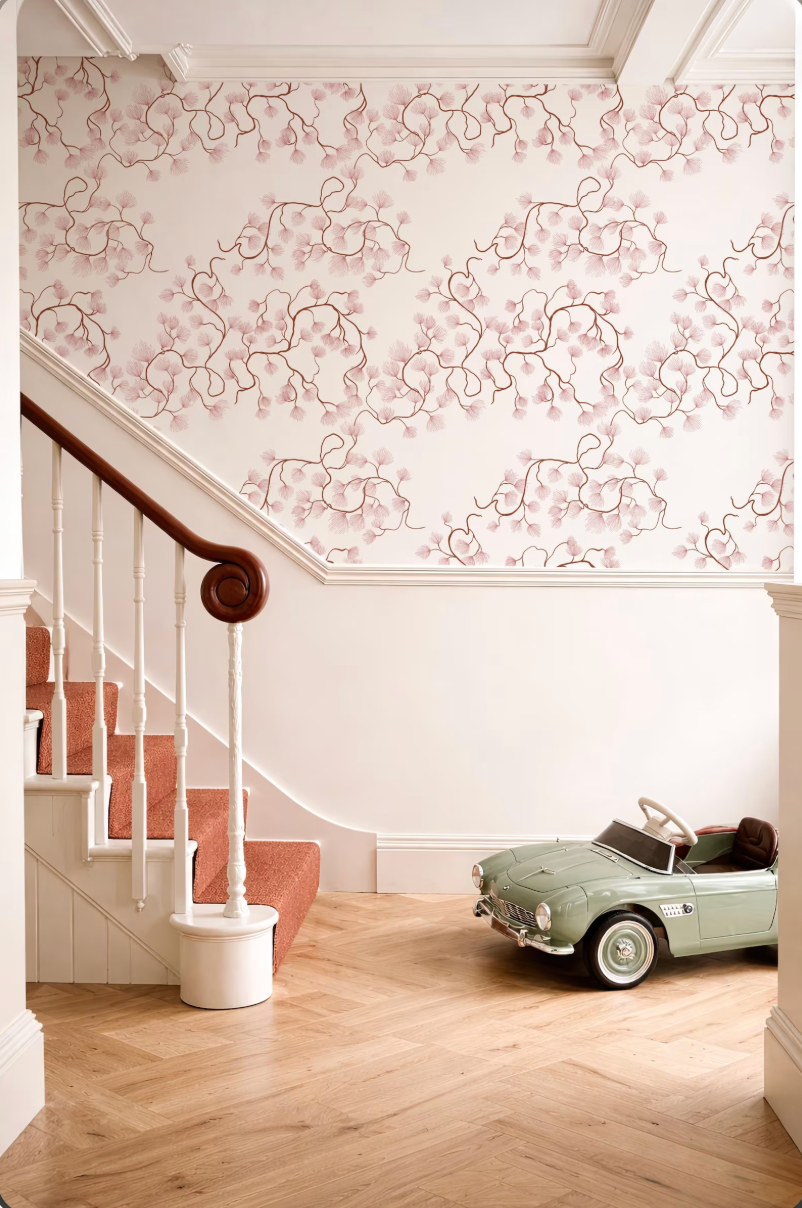 a vintage green toy car parked on a wooden floor next to a white staircase with a wooden handrail. The walls are decorated with a floral pattern, and the overall color scheme is predominantly pink and white.