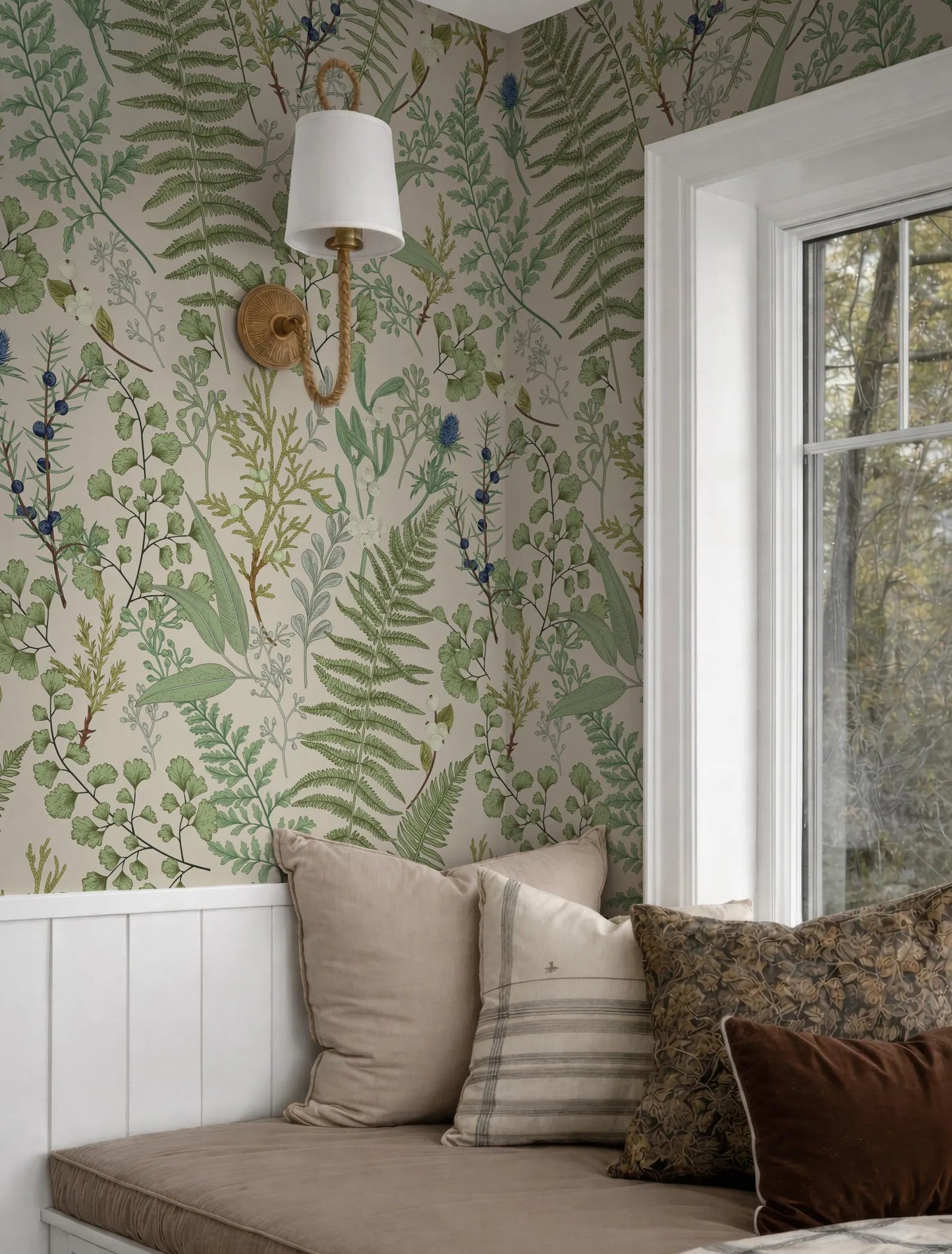 a cozy corner of a room with a large window, a wallpapered wall with a green and white fern pattern, and a white lamp mounted on the wall. The room also contains a bed with a brown and white striped pillow and a brown throw pillow.