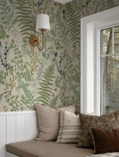 a cozy corner of a room with a large window, a wallpapered wall with a green and white fern pattern, and a white lamp mounted on the wall. The room also contains a bed with a brown and white striped pillow and a brown throw pillow.