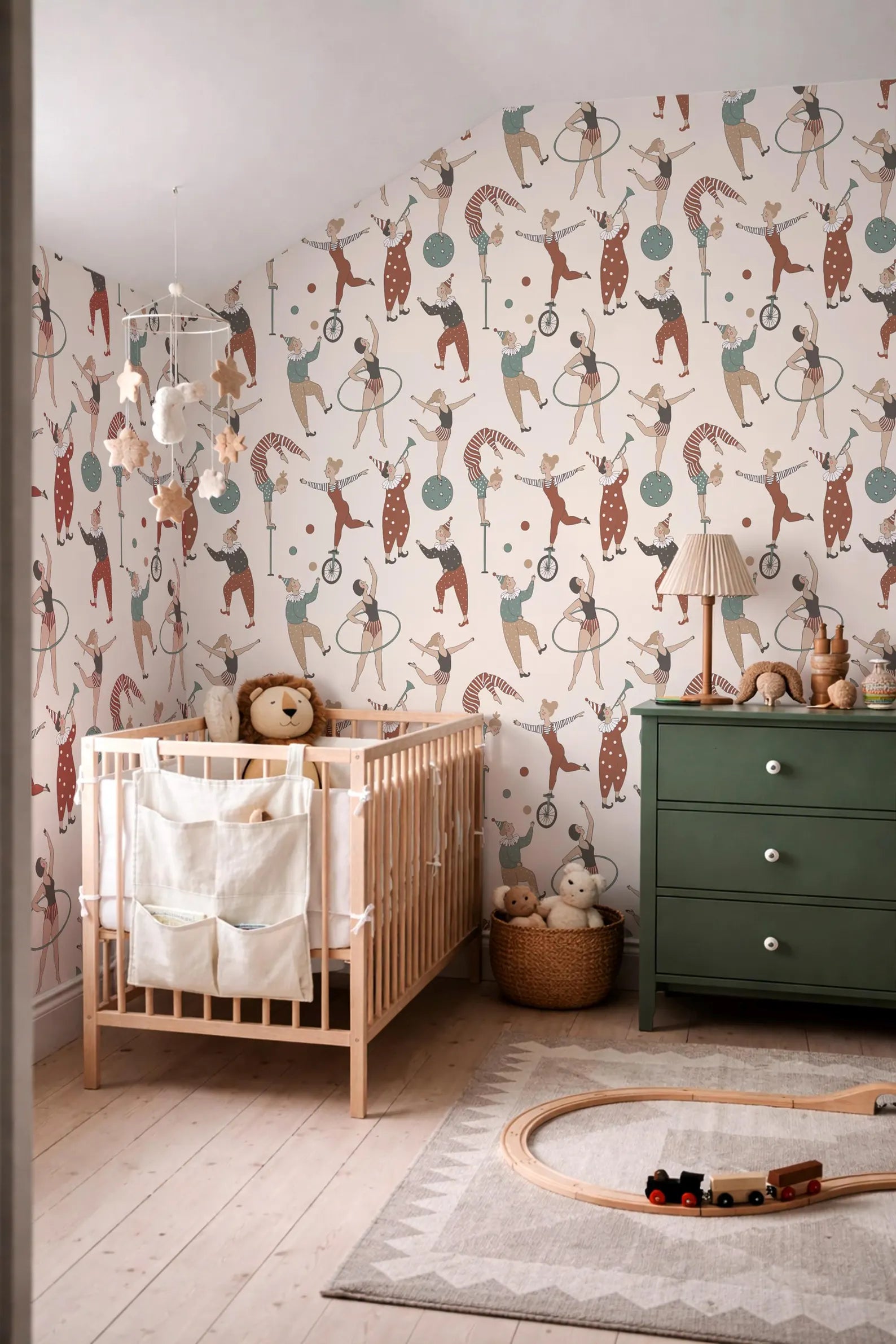 a nursery room with a wooden crib, a green dresser, and a toy train on a rug. The walls are decorated with a pattern of circus performers and circus-related imagery.