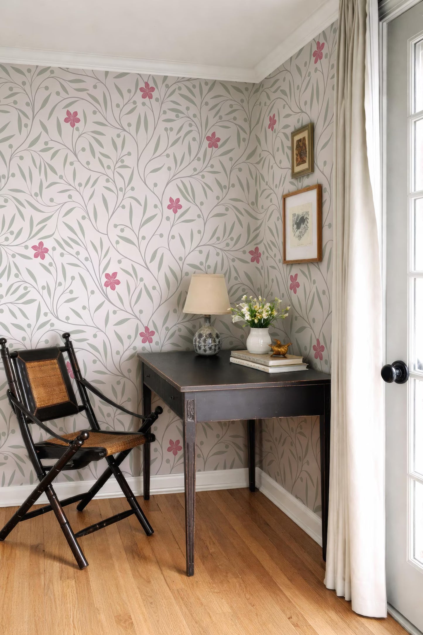 a cozy corner of a room with a wooden desk, a black chair, and a lamp. The walls are adorned with a floral wallpaper, and there is a framed picture hanging above the desk.