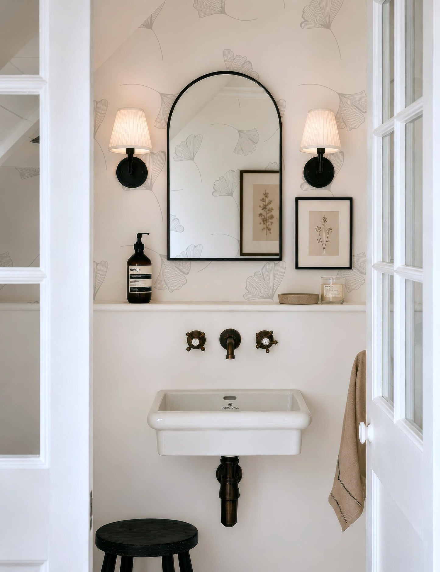 a small bathroom with a white sink, a mirror, and a stool. The walls are decorated with a floral pattern, and there are two wall sconces above the mirror.