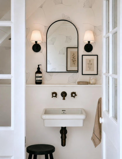 a small bathroom with a white sink, a mirror, and a stool. The walls are decorated with a floral pattern, and there are two wall sconces above the mirror.