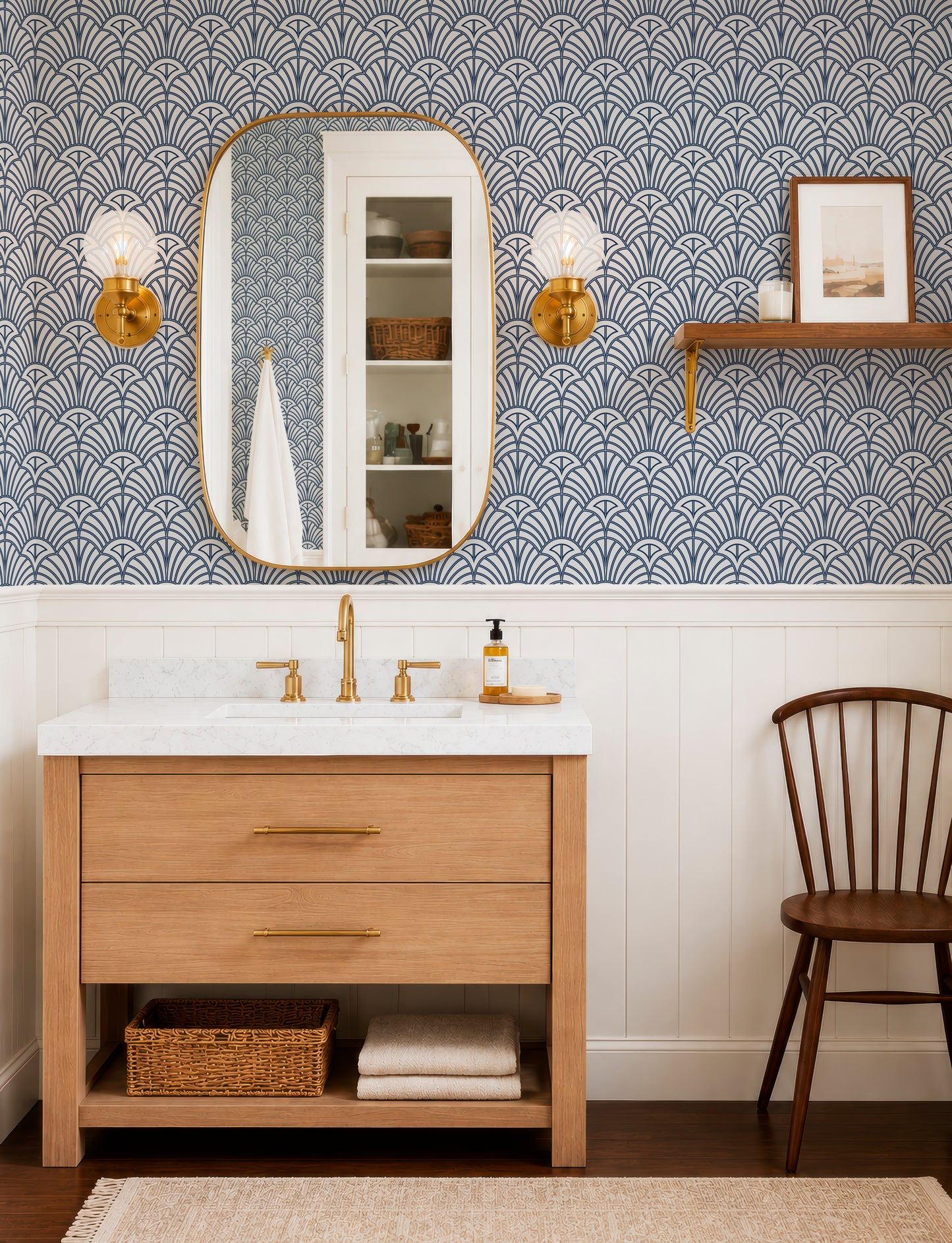 a bathroom with a white sink, a wooden vanity, and a mirror. The walls are covered in a blue and white patterned wallpaper, and there are two gold-framed light fixtures above the mirror. A wooden chair is placed next to the vanity, and a basket is located under the sink.