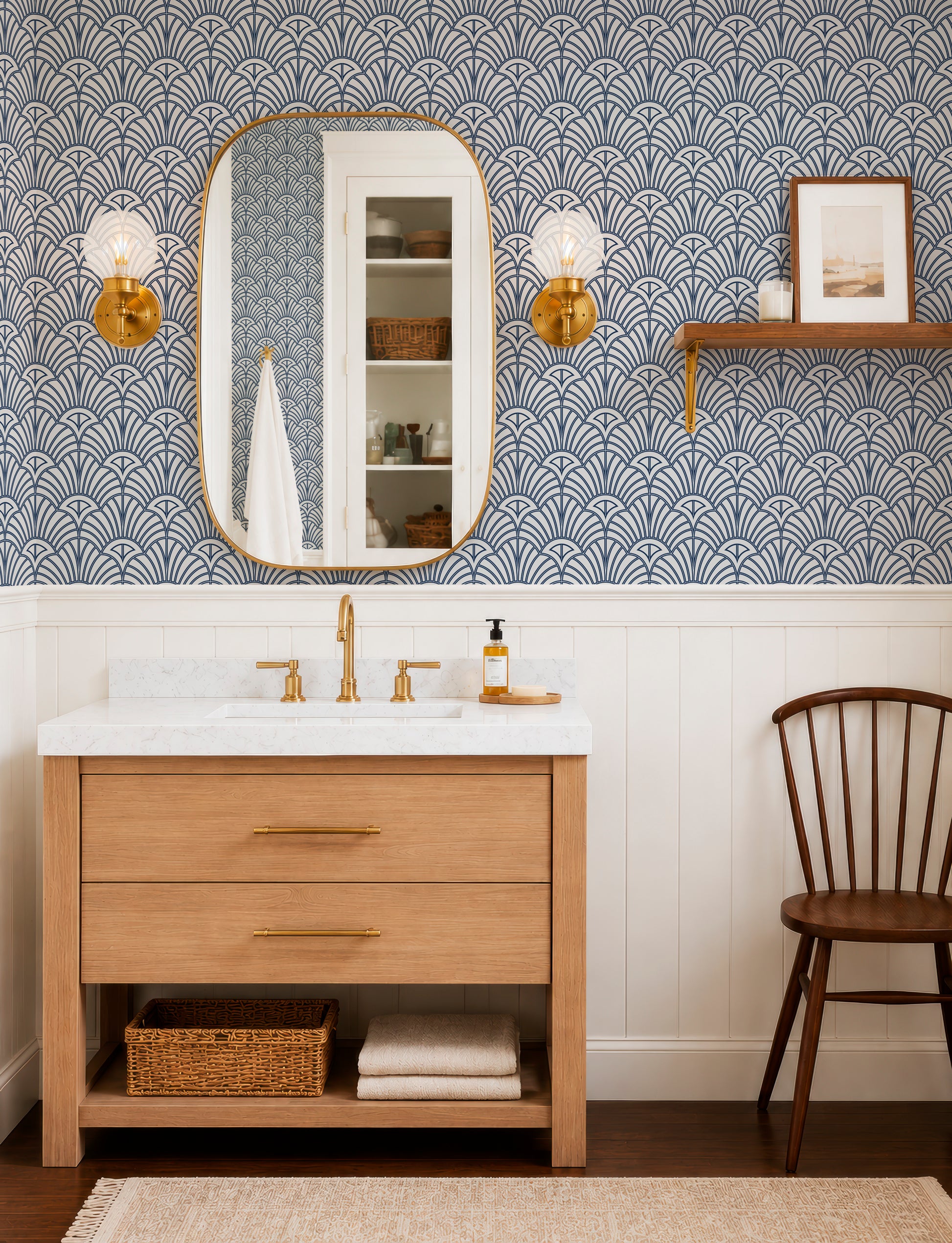 a bathroom with a white sink, a wooden vanity, and a mirror. The walls are covered in a blue and white patterned wallpaper, and there are two gold-framed light fixtures above the mirror. A wooden chair is placed next to the vanity, and a basket is located under the sink.