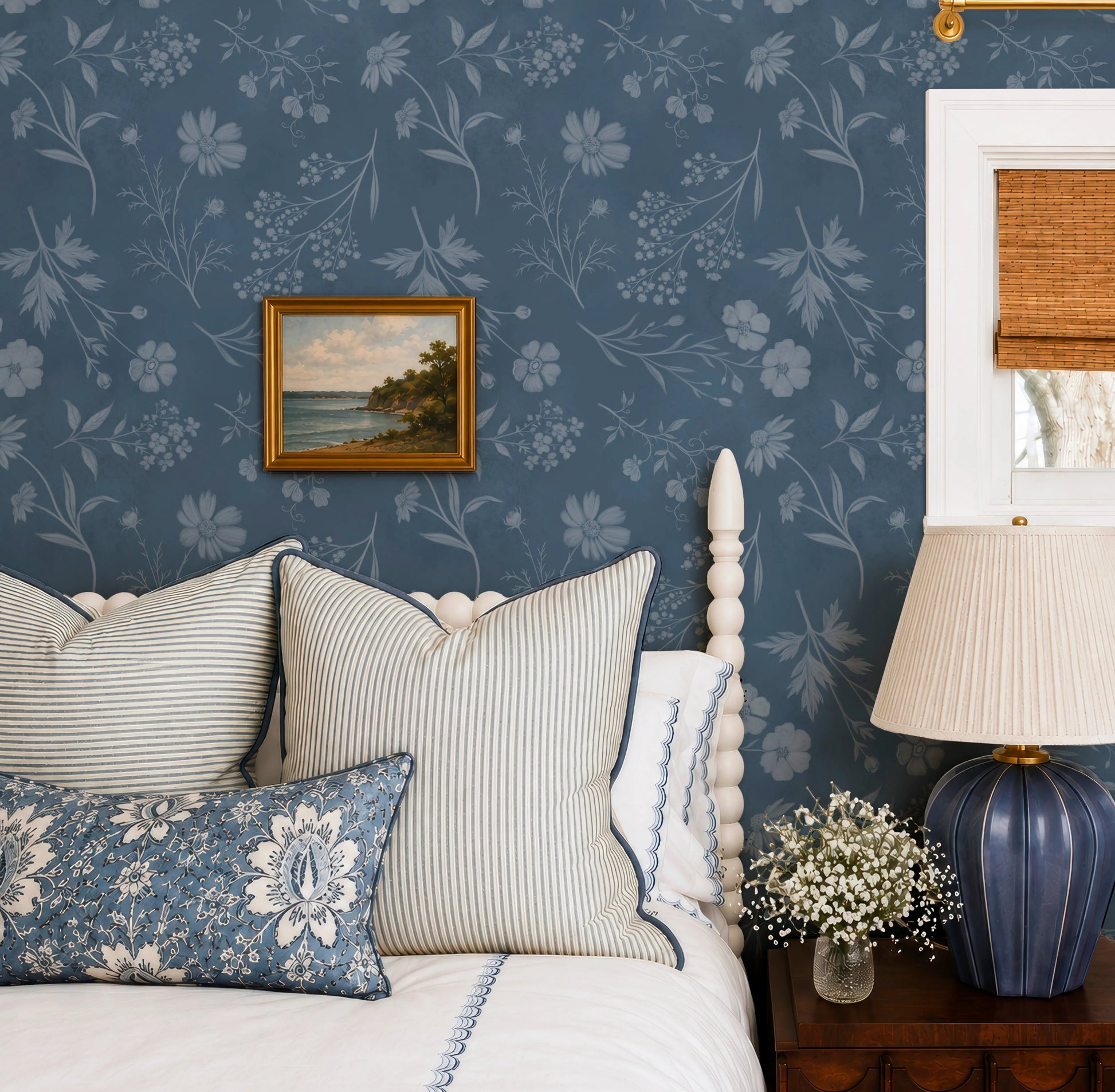 a bedroom with a blue floral wallpaper, a white bed with striped pillows, a blue lamp, and a framed landscape painting on the wall.
