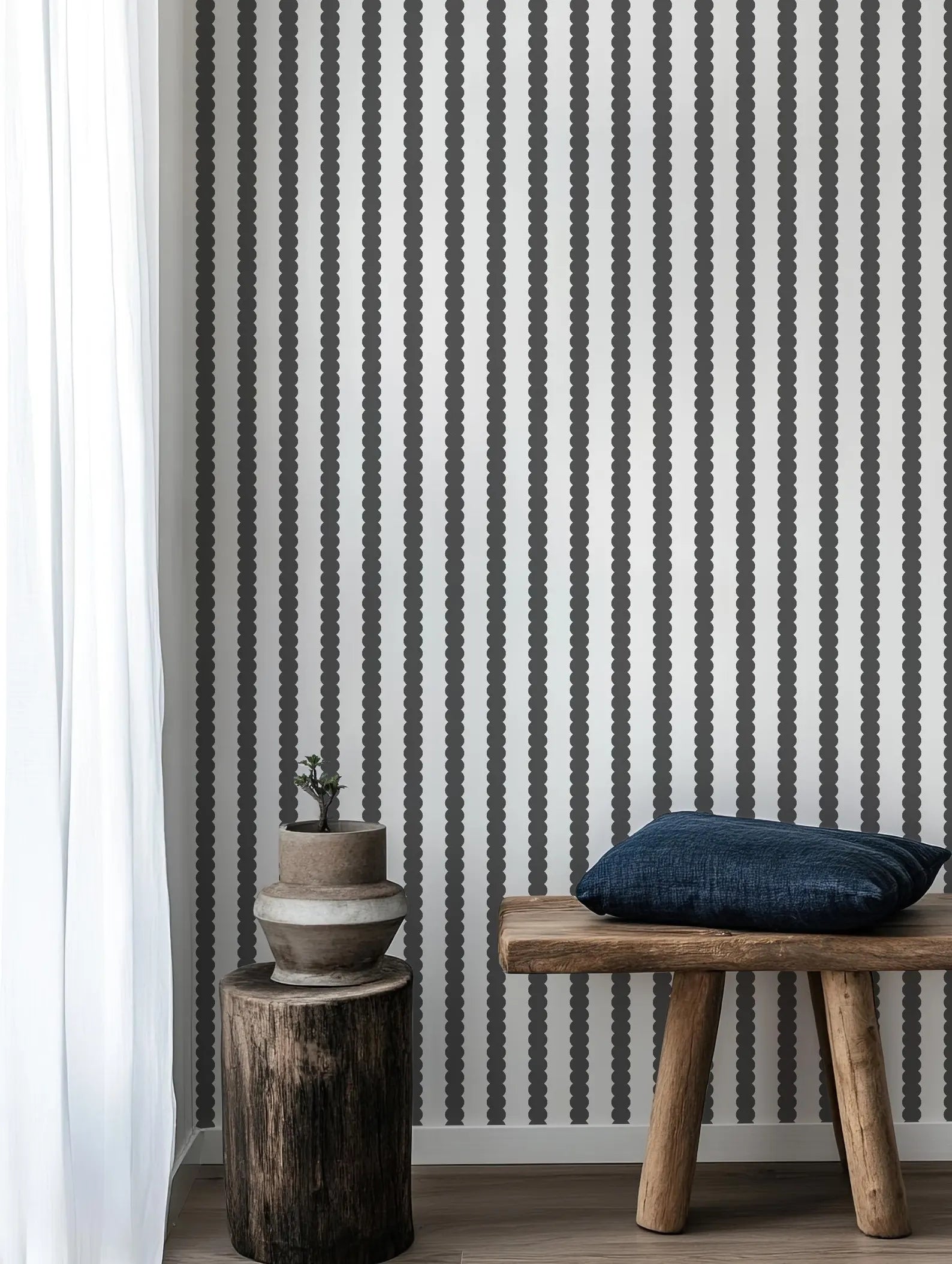 a wooden bench with a blue pillow on it, placed next to a wooden stool with a plant on top. The backdrop is a wall with vertical black and white stripes.
