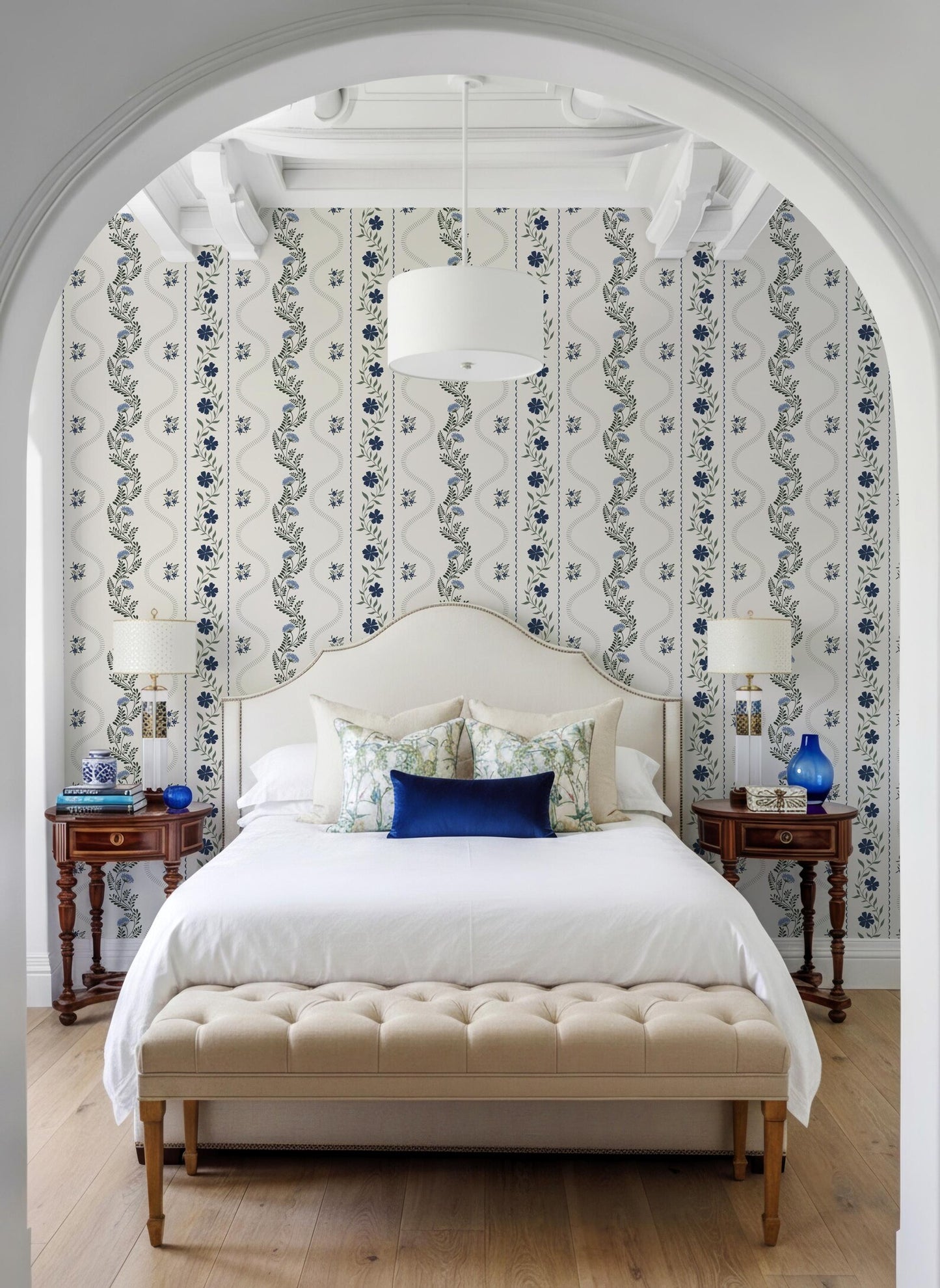 a well-decorated bedroom with a large bed, a wooden nightstand on each side, and a tufted bench in front of the bed. The room has a white archway and a patterned wallpaper, creating a cozy and inviting atmosphere.