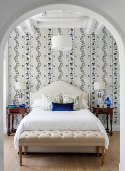 a well-decorated bedroom with a large bed, a wooden nightstand on each side, and a tufted bench in front of the bed. The room has a white archway and a patterned wallpaper, creating a cozy and inviting atmosphere.