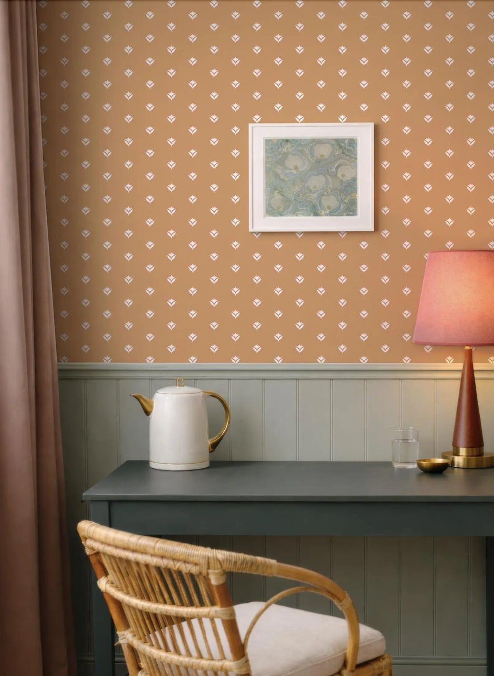 a cozy corner of a room with a wooden desk, a white teapot, and a pink lamp. The wall behind the desk has a patterned wallpaper with small white dots. A framed picture is hanging on the wall above the desk.