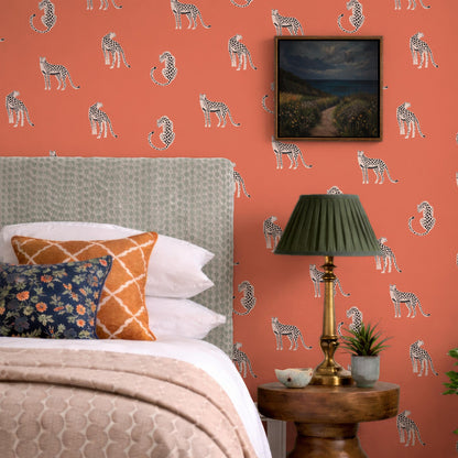 a bedroom with a bed, a lamp, and a framed picture on the wall. The wall has a vibrant orange and white pattern, and the bed is adorned with a floral patterned pillow and a striped pillow. The lamp has a green shade and is placed on a wooden side table. The framed picture on the wall depicts a serene landscape with a winding path and a body of water.