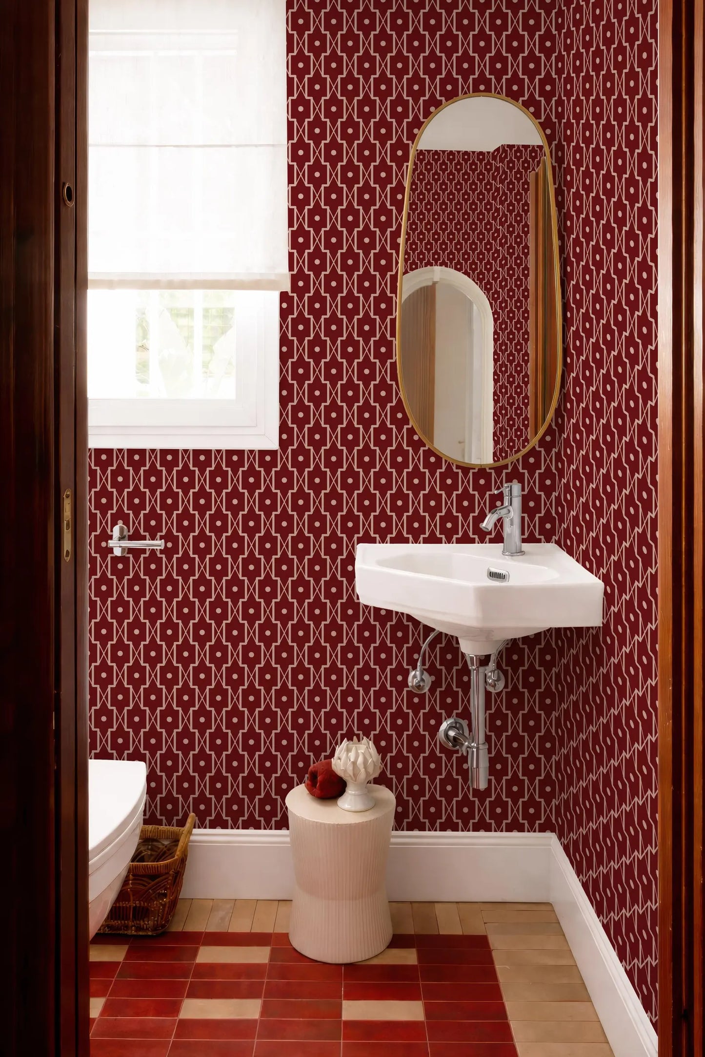a bathroom with a white sink, a mirror, and a window. The walls are covered in a red and white patterned wallpaper, and the floor is made of red and white checkered tiles. There is a small stool in front of the sink, and a basket is placed on the floor next to the sink.