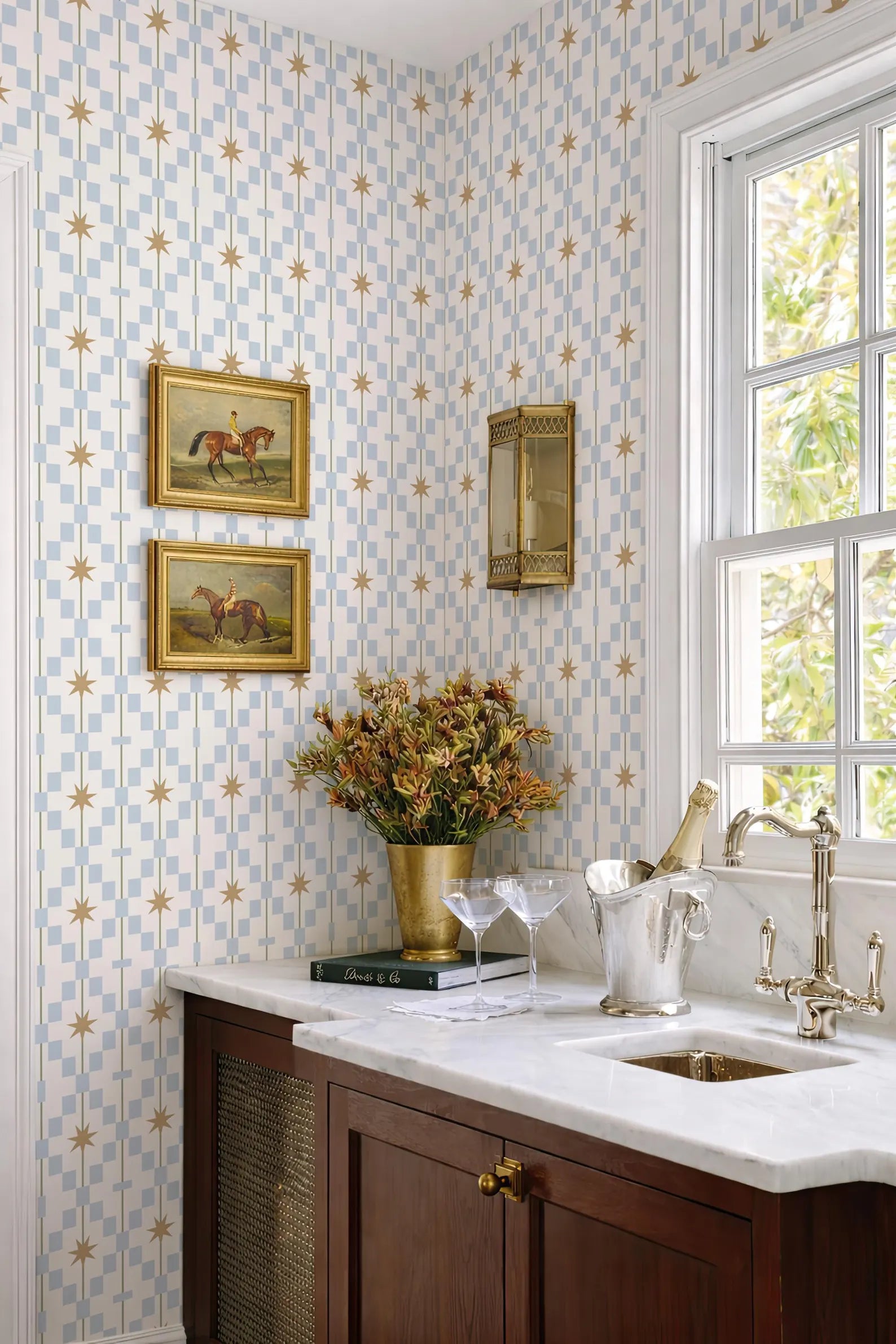 a well-decorated kitchen with a white countertop, a vase of flowers, and a set of wine glasses. The wall behind the countertop has a patterned wallpaper with blue and white stars. There are two framed pictures hanging on the wall, and a window with white curtains allows natural light to enter the space.