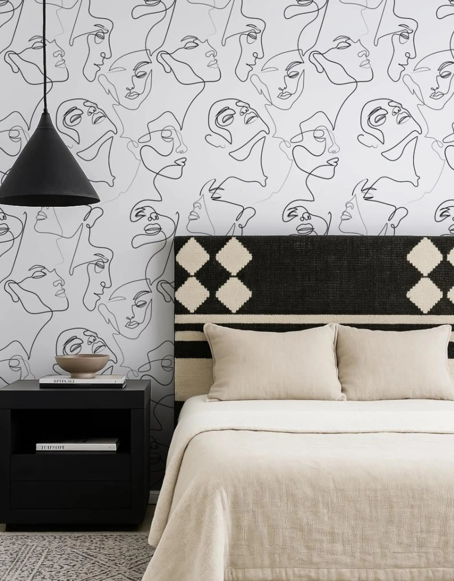 a bedroom with a black and white abstract wallpaper pattern on the wall behind a bed with a black and white patterned headboard. The bed is dressed in a beige comforter and has two pillows. A black nightstand is positioned next to the bed, and a black pendant light hangs above it.