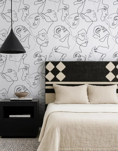 a bedroom with a black and white abstract wallpaper pattern on the wall behind a bed with a black and white patterned headboard. The bed is dressed in a beige comforter and has two pillows. A black nightstand is positioned next to the bed, and a black pendant light hangs above it.