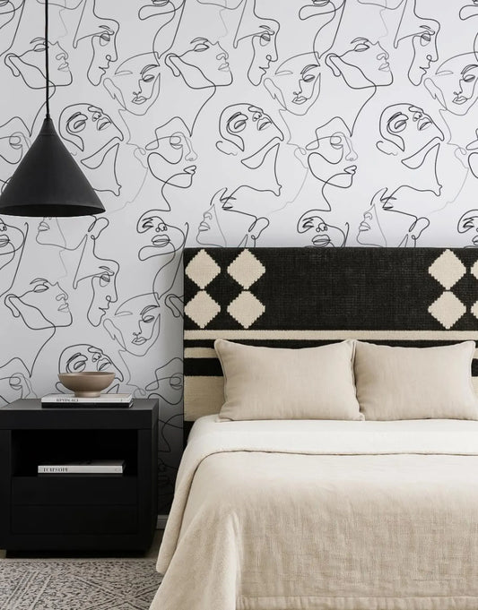 a bedroom with a black and white abstract wallpaper pattern on the wall behind a bed with a black and white patterned headboard. The bed is dressed in a beige comforter and has two pillows. A black nightstand is positioned next to the bed, and a black pendant light hangs above it.