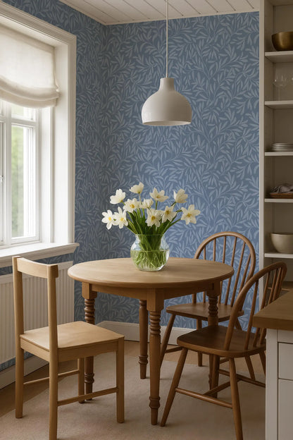 a cozy dining area with a wooden table, two chairs, and a vase of white flowers on the table. The room has a blue and white patterned wallpaper, and a white pendant light hangs above the table.