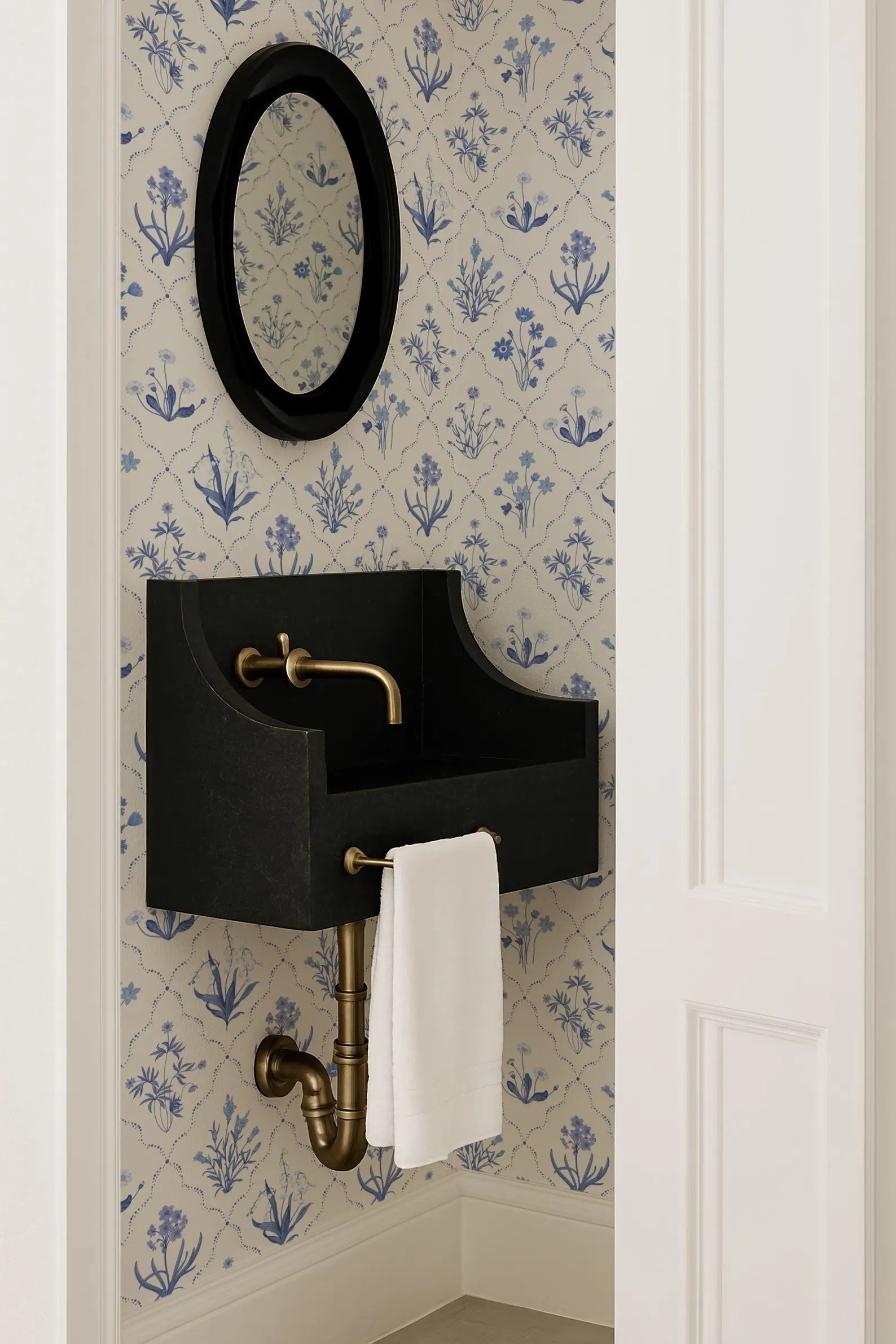 a bathroom with a black sink and a white towel hanging on a pipe. The sink is mounted on the wall, and there is a mirror above it. The wall has a blue and white floral pattern.