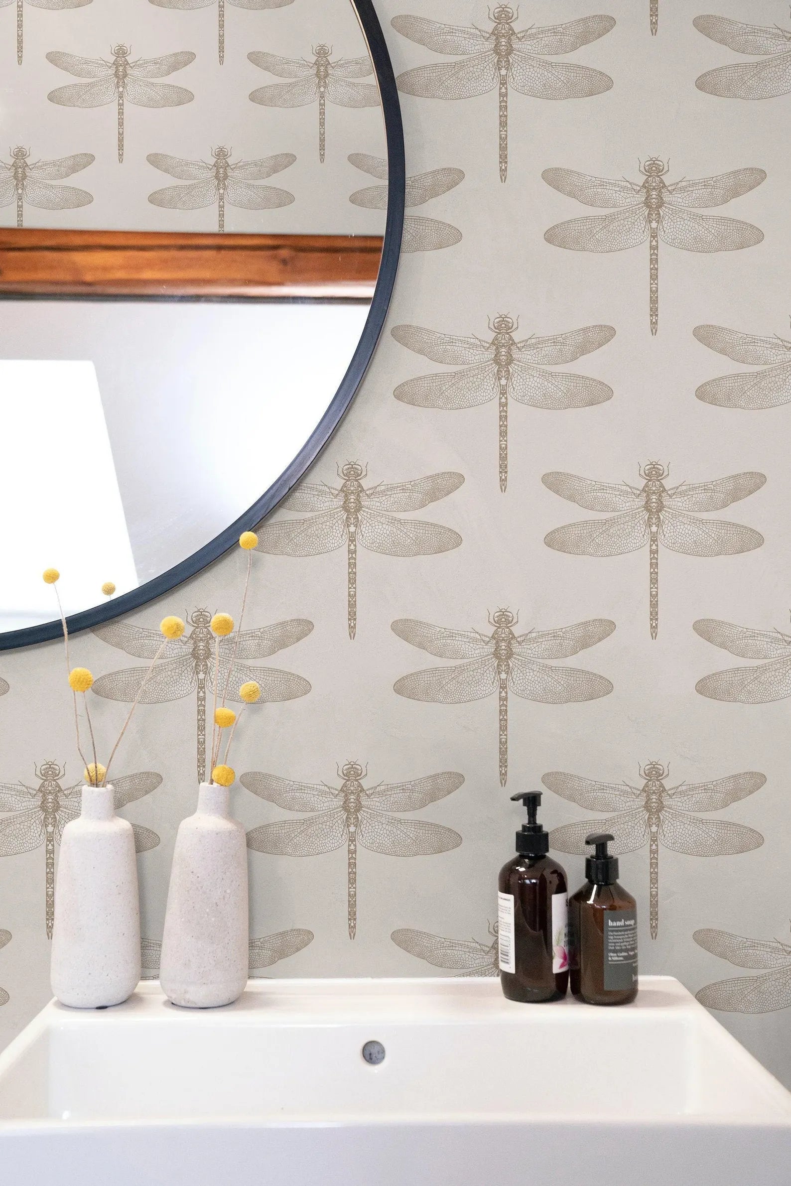 a bathroom setting with a white sink, a round mirror, and a wallpapered wall with a repeating pattern of dragonflies. There are two white vases with yellow flowers on the counter, and two bottles of soap are placed next to the sink.
