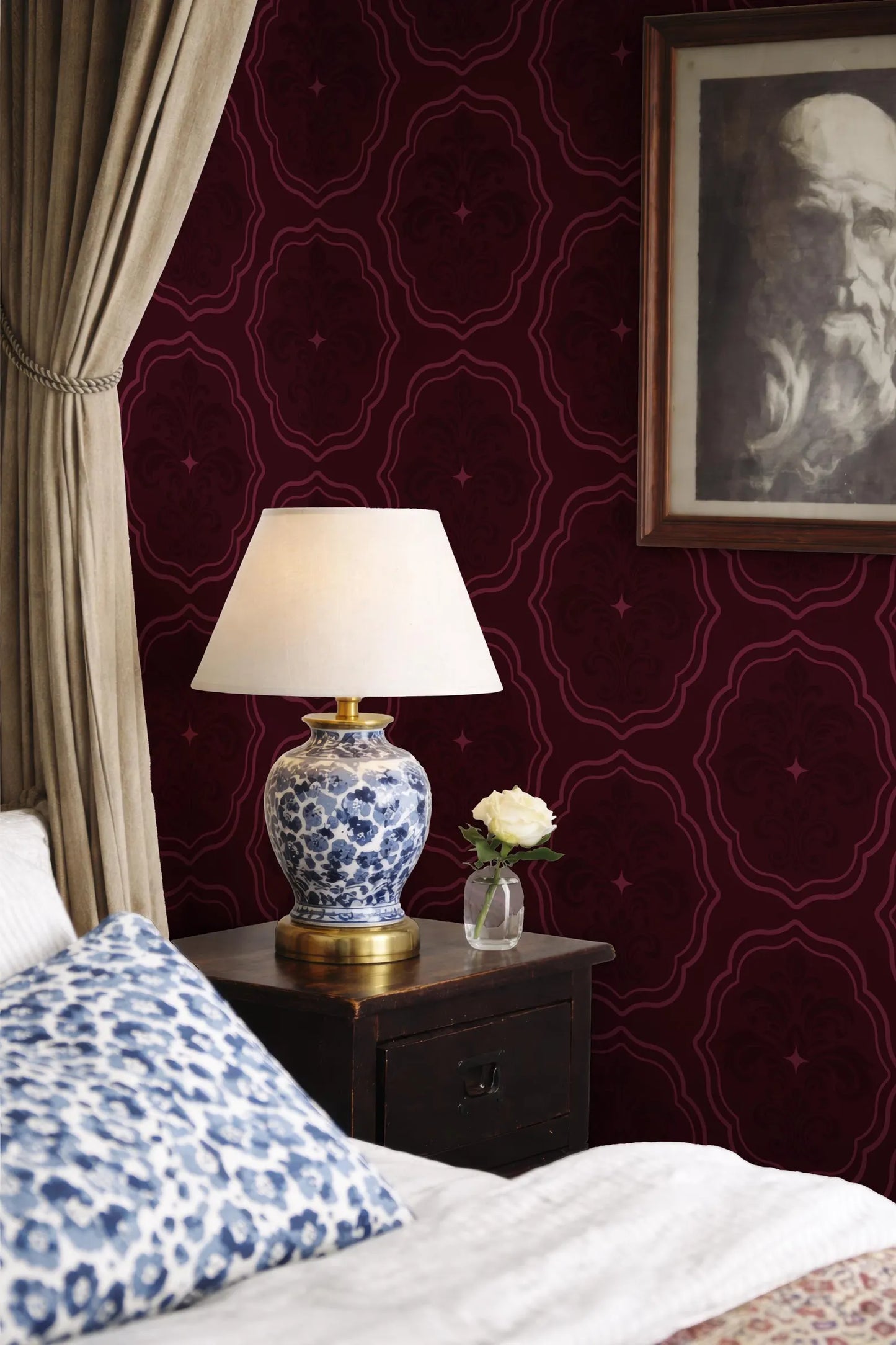 a bedroom with a bed, a nightstand, and a lamp with a white shade. The wall behind the nightstand has a red and burgundy patterned wallpaper. A framed picture is hanging on the wall, and a vase with a single white rose is placed on the nightstand.