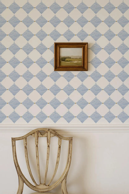 a white chair with a wooden frame, positioned against a wall with a diamond pattern wallpaper. A small framed landscape painting is hanging on the wall above the chair.