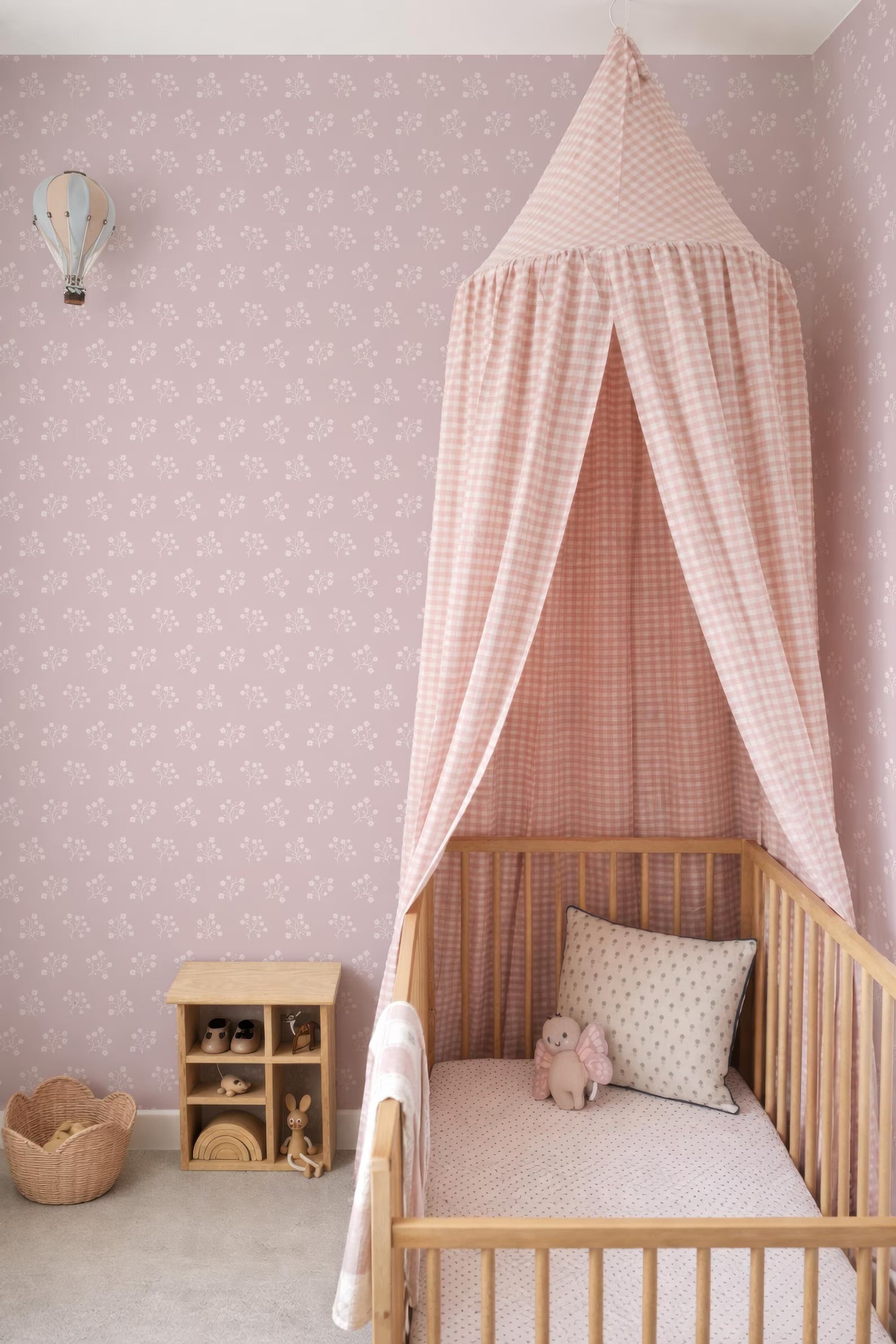 A cozy nursery with a wooden crib, a canopy bed, and a small wooden shelf with toys. The walls are decorated with pink floral wallpaper, and a small basket is placed on the floor.