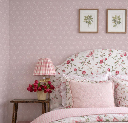 a bedroom with a floral-patterned headboard, a lamp with a pink and white striped shade, and a bed with matching bedding. The walls are covered in pink wallpaper with a floral pattern, and there are two framed botanical illustrations on the wall above the bed.
