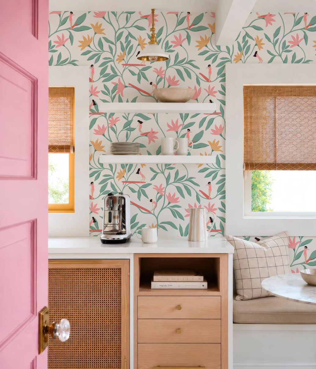 a kitchen with a pink door, a white countertop, and a wooden cabinet. The wall behind the countertop has a floral pattern wallpaper. On the countertop, there is a coffee maker, a stack of books, and a bowl.