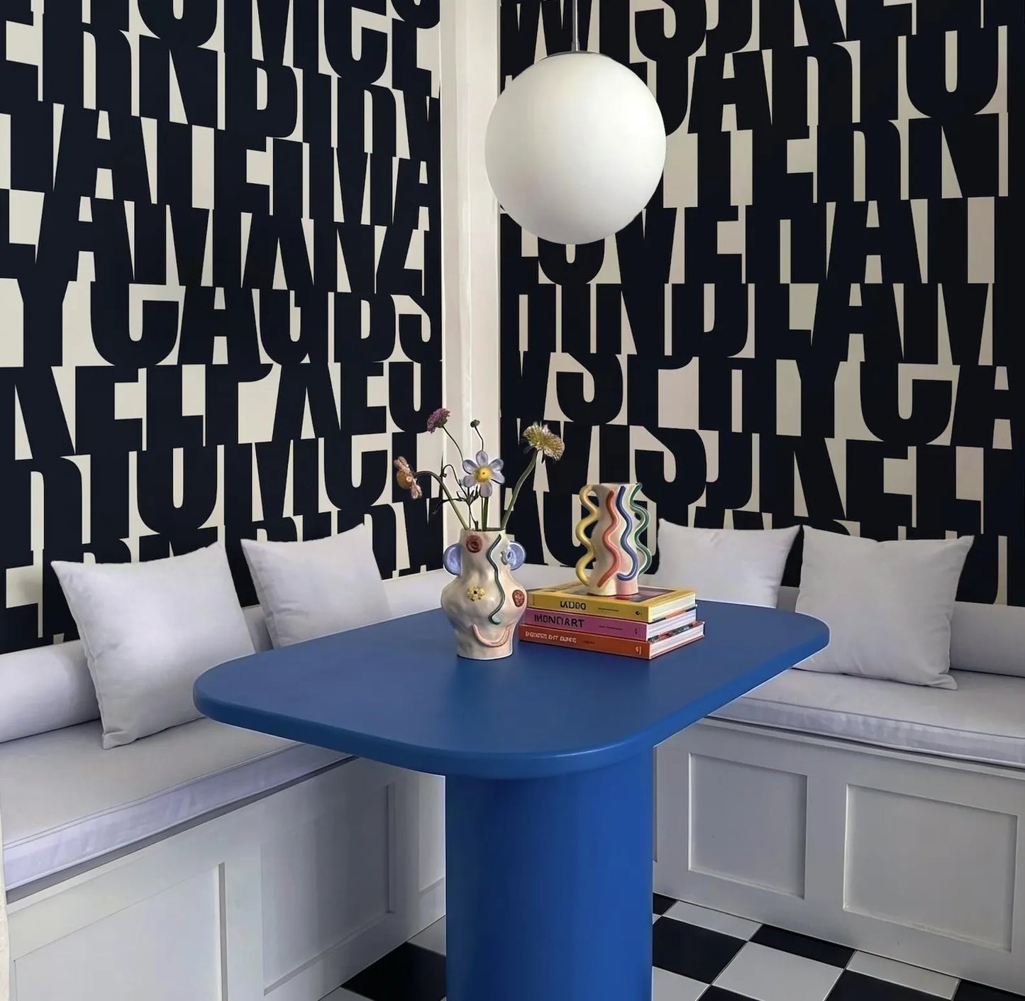 a modern and stylish dining area with a blue table, white cushions, and a black and white checkered floor. The wall behind the table is covered in large black letters, creating a bold and striking visual effect. On the table, there is a vase with flowers and a stack of books, adding a touch of color and personality to the space.