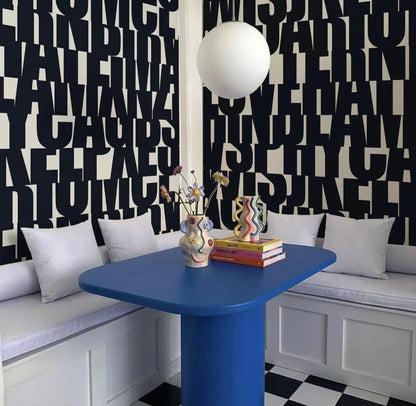 a modern and stylish dining area with a blue table, white cushions, and a black and white checkered floor. The wall behind the table is covered in large black letters, creating a bold and striking visual effect. On the table, there is a vase with flowers and a stack of books, adding a touch of color and personality to the space.