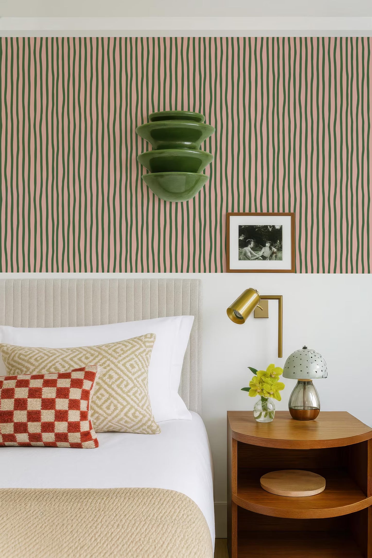 a well-decorated bedroom with a bed, nightstand, and wall art. The bed has a white comforter and a red and white checkered pillow. The nightstand has a lamp and a vase with yellow flowers. The wall behind the bed has a striped wallpaper pattern. There is also a framed photo hanging on the wall.