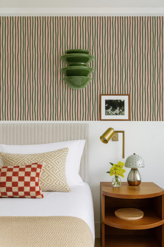 a well-decorated bedroom with a bed, nightstand, and wall art. The bed has a white comforter and a red and white checkered pillow. The nightstand has a lamp and a vase with yellow flowers. The wall behind the bed has a striped wallpaper pattern. There is also a framed photo hanging on the wall.