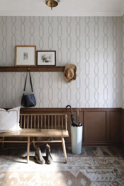 a cozy entryway with a wooden bench, a shelf with framed pictures, a hat rack, and a pair of boots. The walls are adorned with a patterned wallpaper, and a rug is placed on the floor.