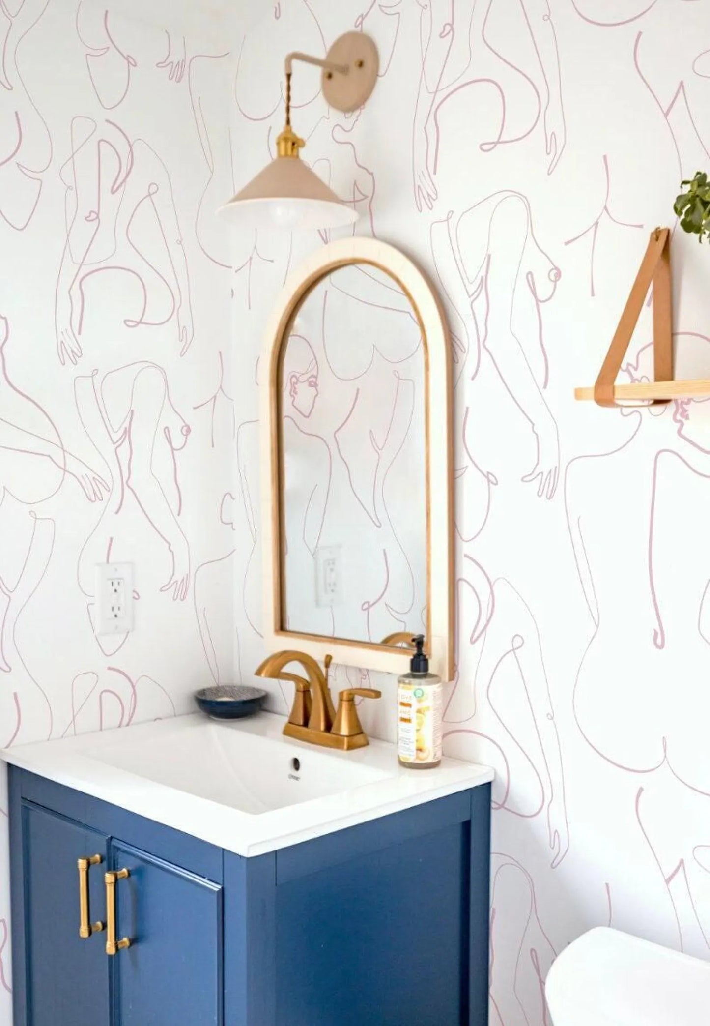 a bathroom with a blue vanity, a gold faucet, and a mirror with a gold frame. The walls are decorated with a pink and white abstract pattern, and there is a small plant on the vanity.
