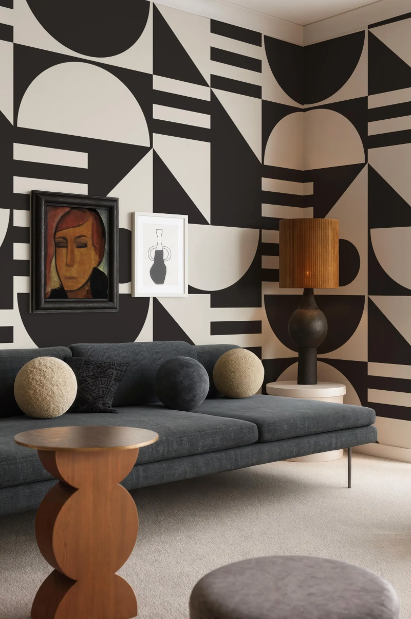 a modern living room with a gray sofa, a wooden side table, and a lamp. The walls are adorned with a geometric pattern in black and white, and there is a framed painting hanging above the sofa.
