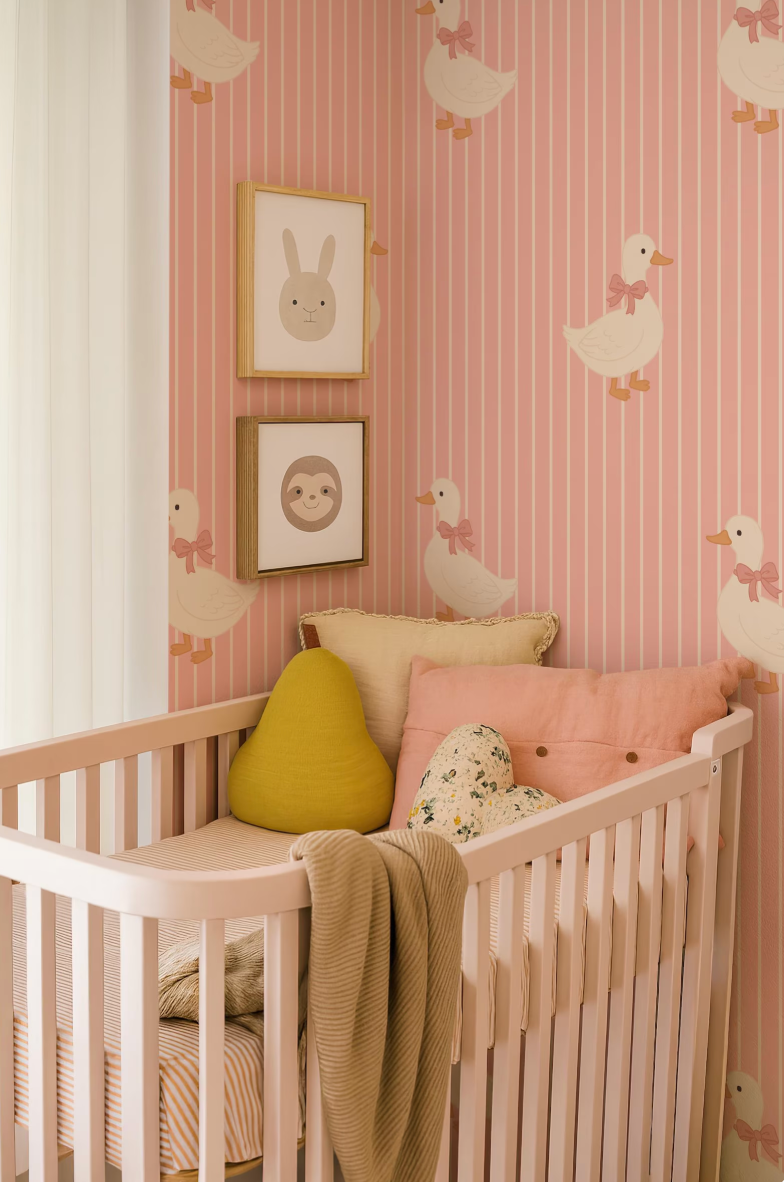 A cozy nursery with a white crib, adorned with a yellow pillow and a pink blanket. The walls are decorated with a pink and white striped wallpaper featuring cartoon duck illustrations. Two framed animal faces are displayed on the wall above the crib.