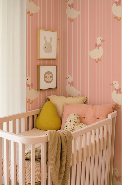 A cozy nursery with a white crib, adorned with a yellow pillow and a pink blanket. The walls are decorated with a pink and white striped wallpaper featuring cartoon duck illustrations. Two framed animal faces are displayed on the wall above the crib.