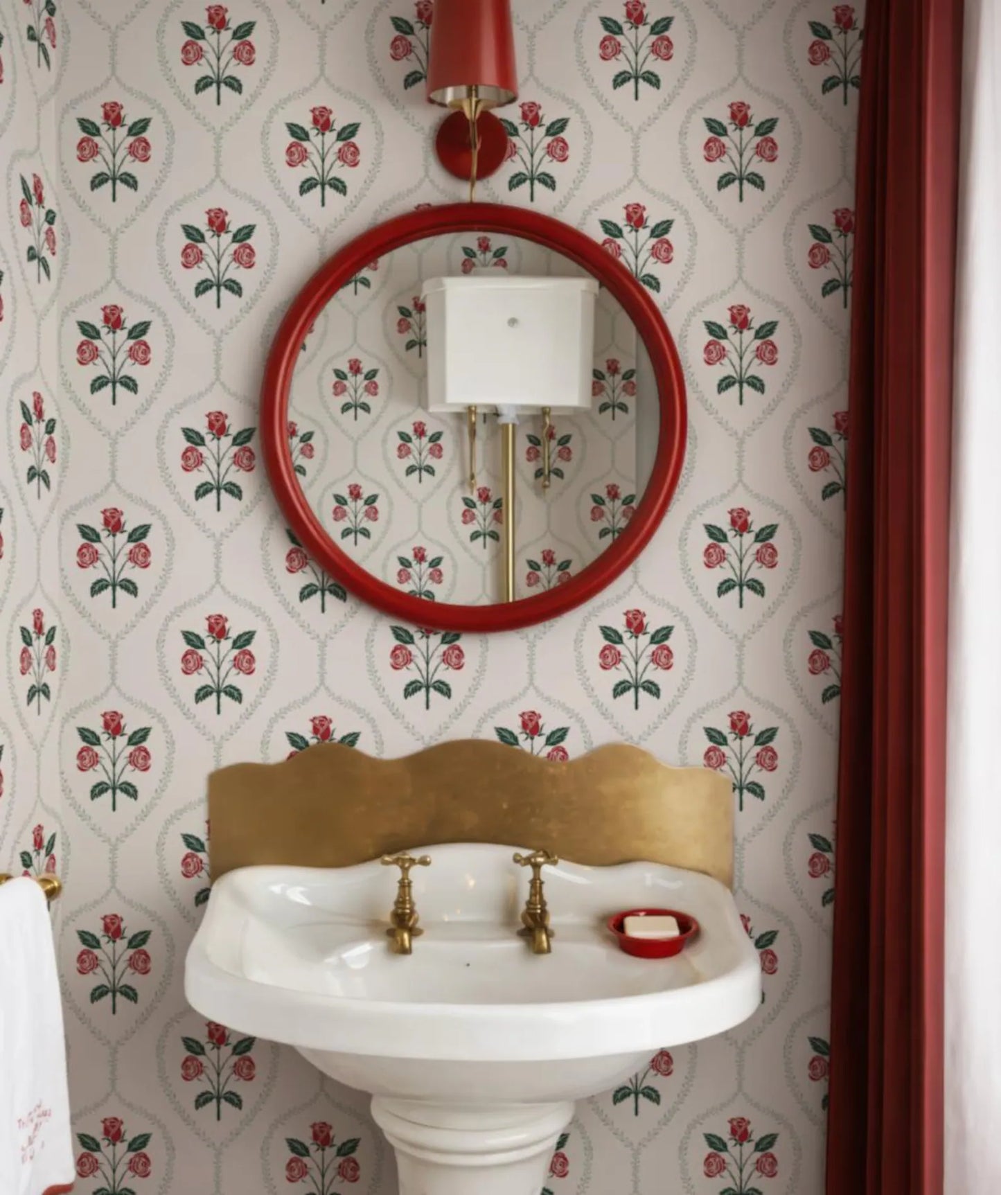 a bathroom with a white pedestal sink, a red-framed mirror, and a floral wallpaper pattern.