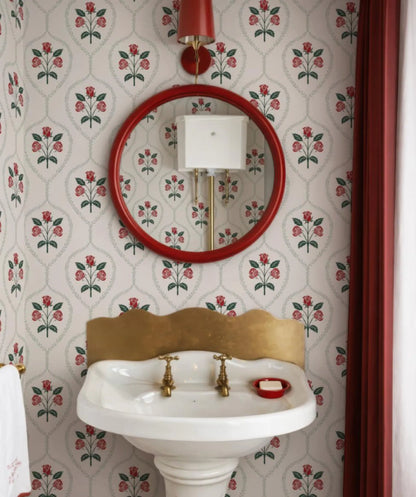 a bathroom with a white pedestal sink, a red-framed mirror, and a floral wallpaper pattern.