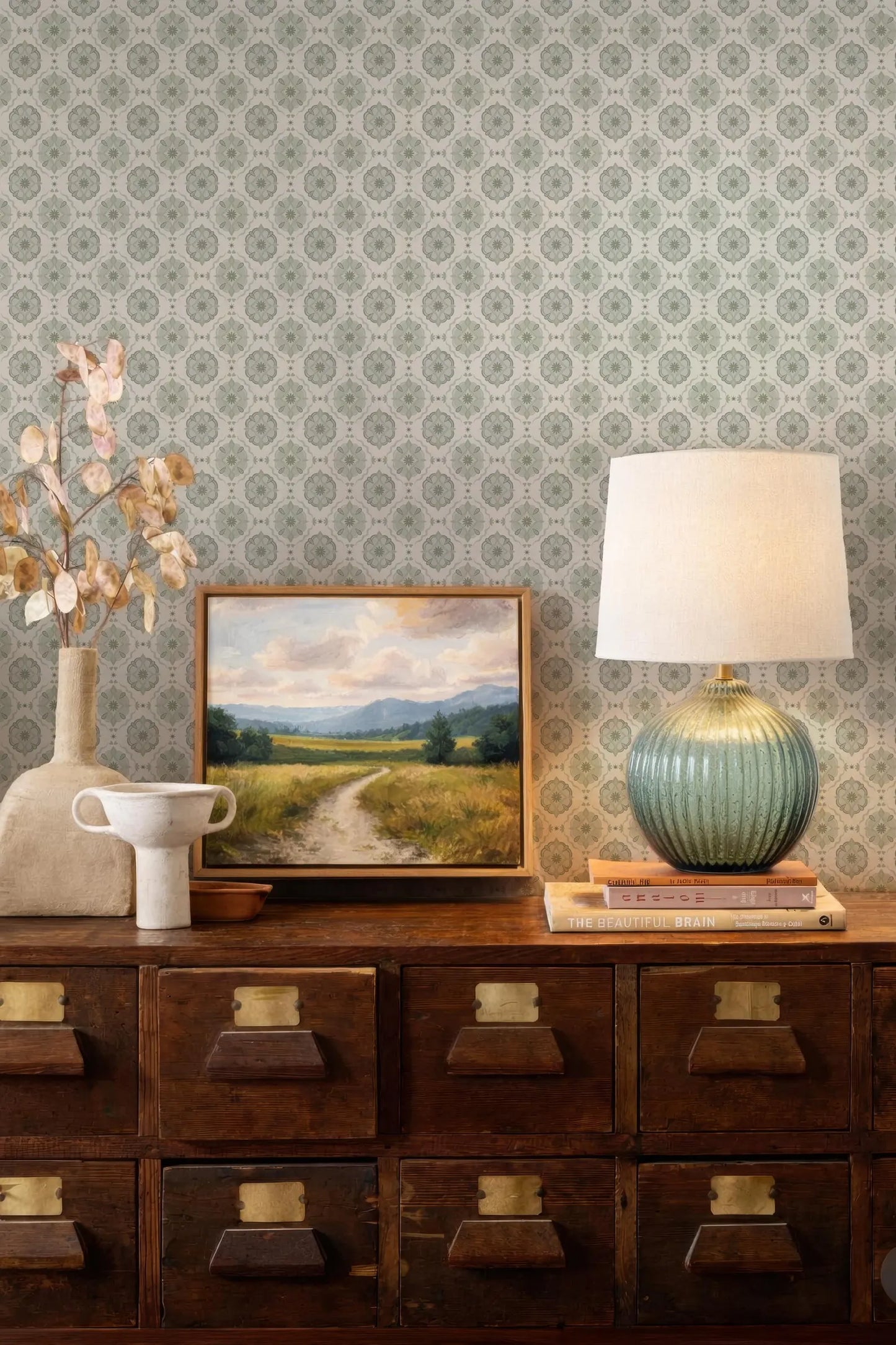 a wooden dresser with a painting, a lamp, and a vase on top. The dresser has multiple drawers and a shelf. The wall behind the dresser has a patterned wallpaper.