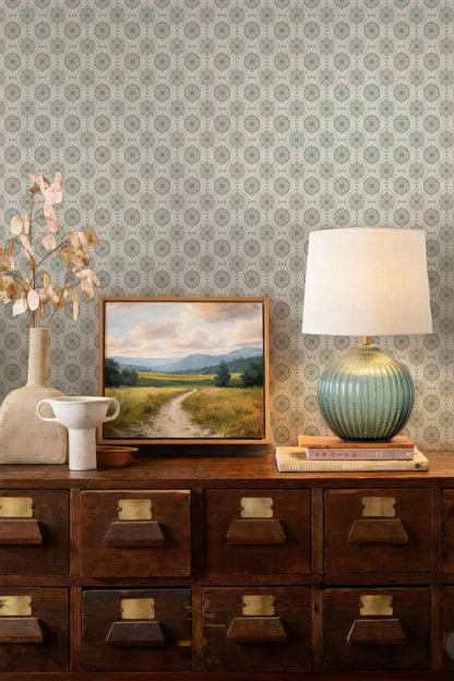a wooden dresser with a painting, a lamp, and a vase on top. The dresser has multiple drawers and a shelf. The wall behind the dresser has a patterned wallpaper.