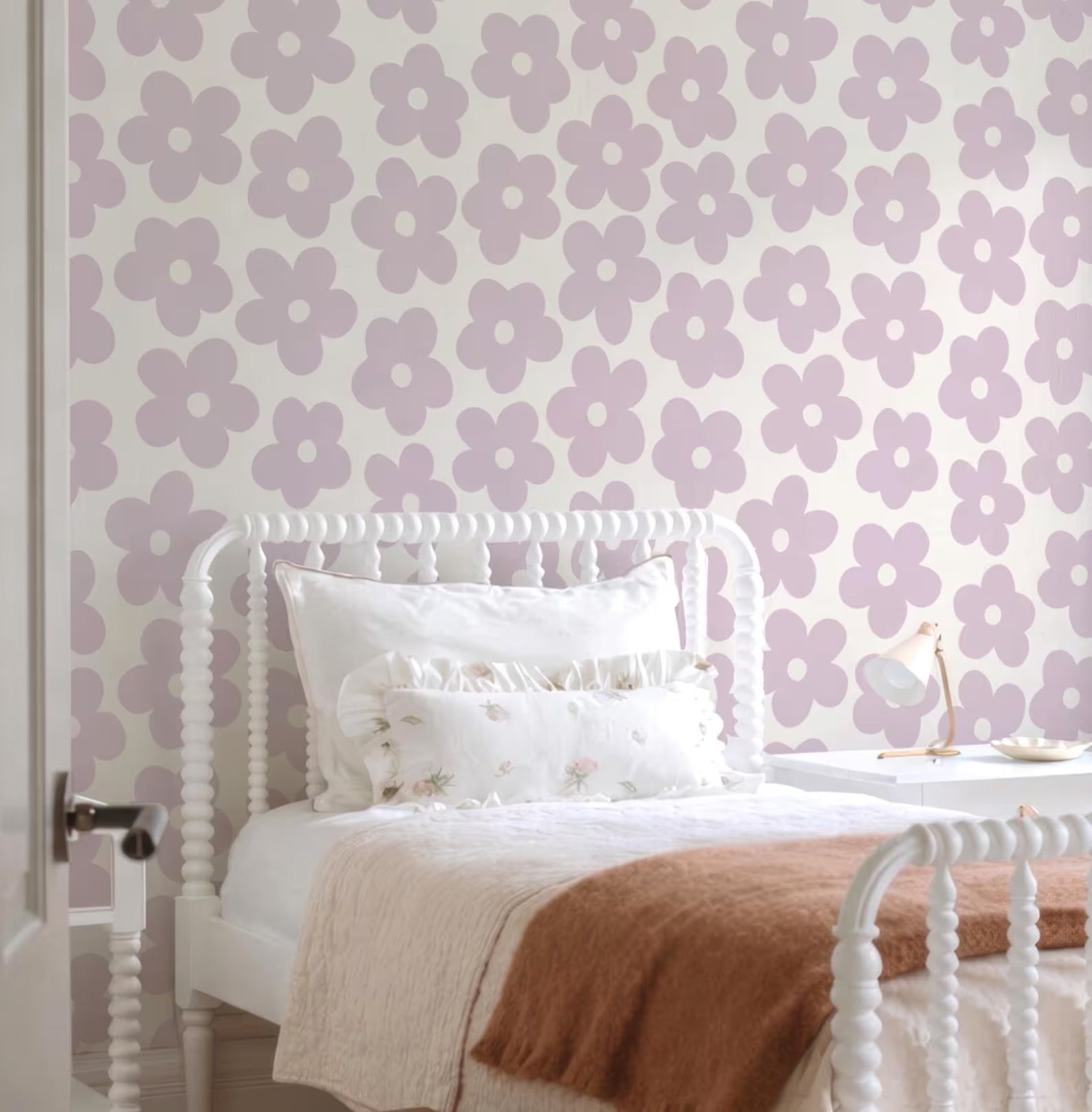 a cozy bedroom with a white bed frame, a white headboard, and a purple and white floral wallpaper. The bed is neatly made with a white comforter and a brown throw blanket. There is a nightstand with a lamp on it, and a small table next to the bed.