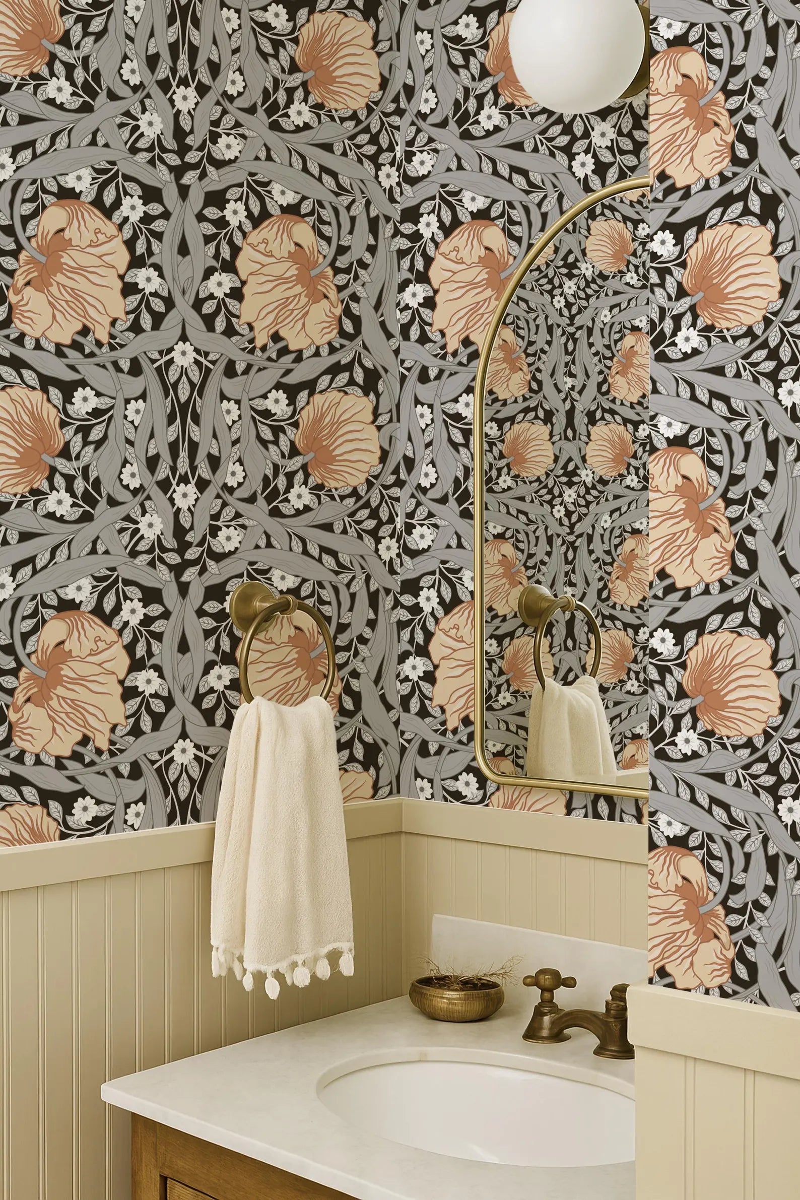 a bathroom with a white sink, a mirror, and a towel rack. The walls are covered in a floral patterned wallpaper, and the overall color scheme is neutral, with a gold-toned metal frame and a white light fixture.