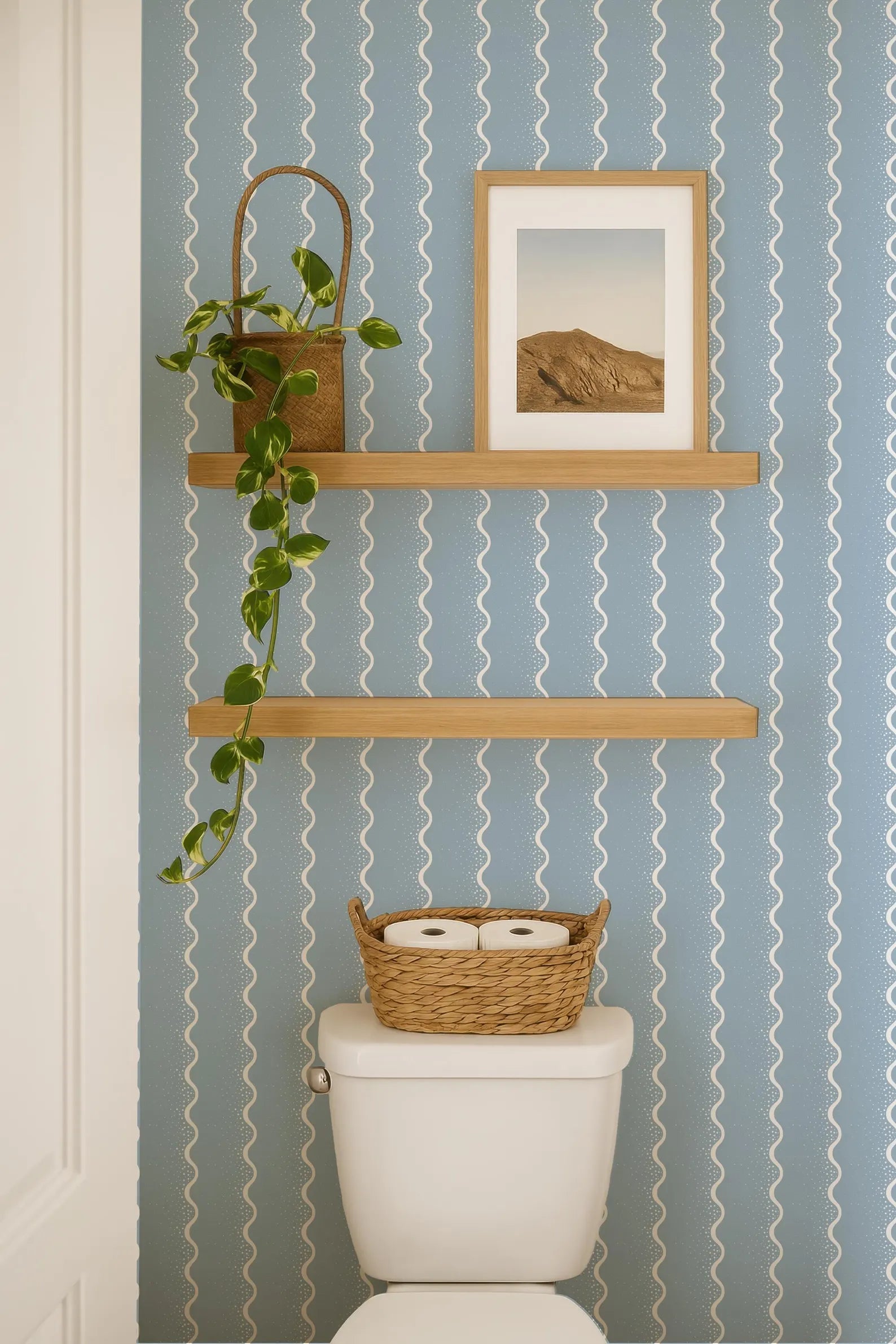 a bathroom with a white toilet, a wooden shelf above it, and a basket of toilet paper on the tank. The wall has a blue and white striped pattern, and there is a framed picture hanging on the wall.