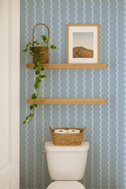 a bathroom with a white toilet, a wooden shelf above it, and a basket of toilet paper on the tank. The wall has a blue and white striped pattern, and there is a framed picture hanging on the wall.