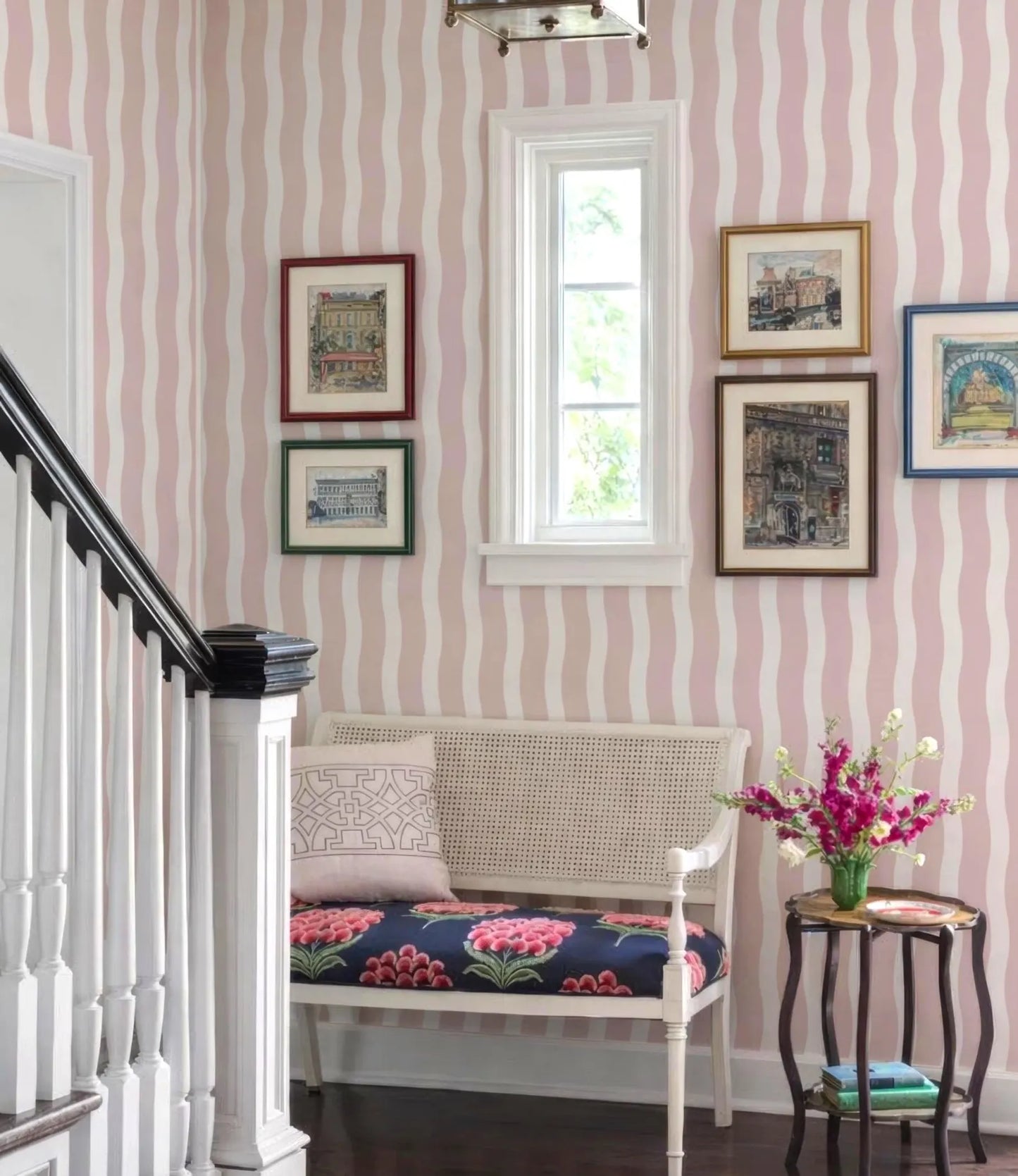 a cozy and charming entryway with a white bench, a small side table with a vase of flowers, and a staircase. The walls are adorned with framed pictures, and there is a window that allows natural light to enter the space.