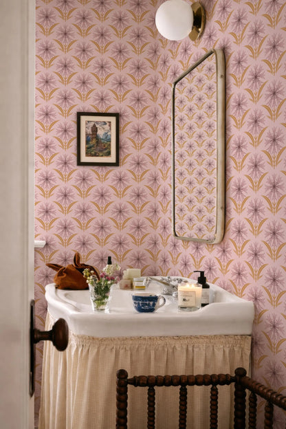 a bathroom with a white sink, a mirror, and a floral patterned wallpaper. The sink is set against a pink and gold floral wallpaper, and there is a vase of flowers on the counter. The mirror is mounted above the sink, and a candle is placed on the counter.