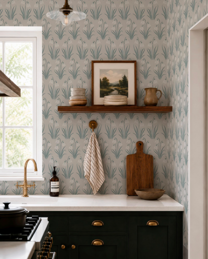 a kitchen with a white countertop, dark cabinets, and a wooden cutting board. Above the countertop, there is a shelf with a framed picture and various kitchen items such as a bowl, a bottle, and a plate. The wall behind the countertop has a patterned wallpaper, and there is a window in the background.