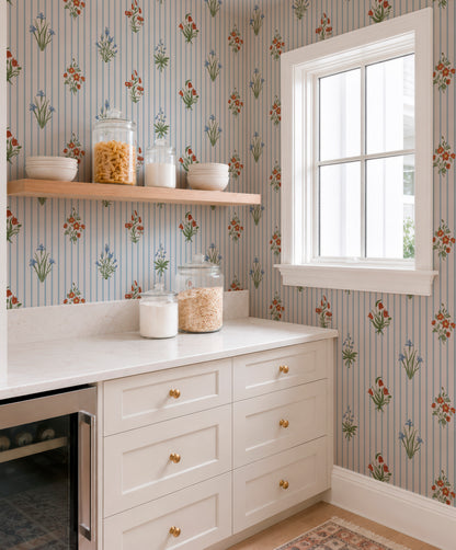 a kitchen with a white countertop, a shelf above it holding various items, and a window with a white frame. The walls are decorated with a floral wallpaper pattern, and the overall color scheme is predominantly white and blue.